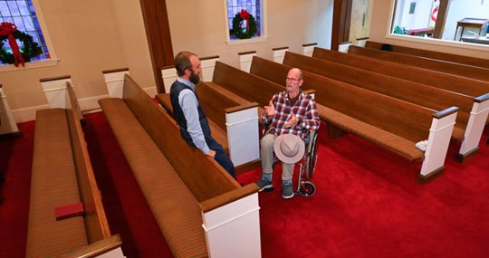 Man in wheelchair talking to pastor in a space with a shorter pew.