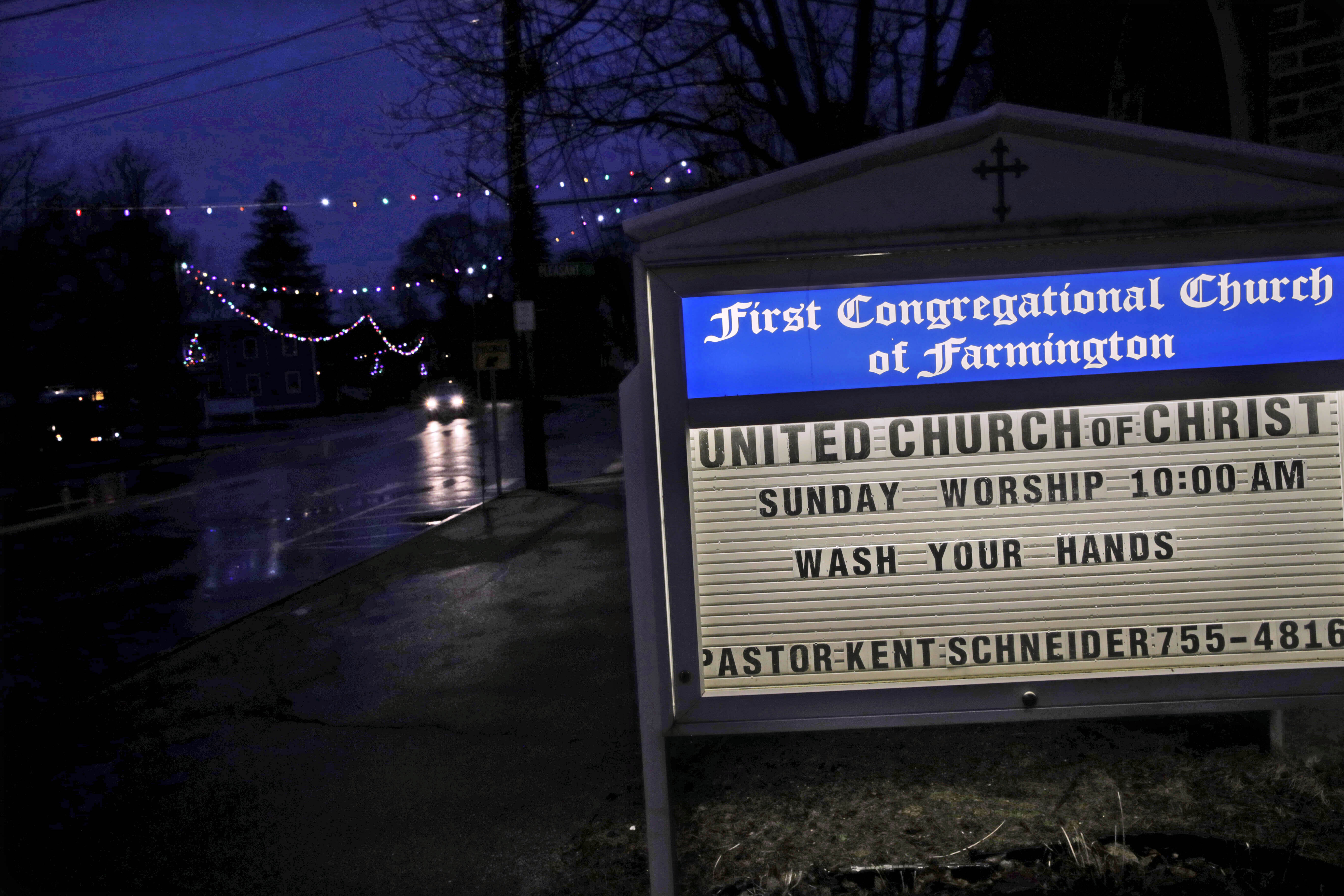 In this March 19, 2020, photo, a church sign reminds townspeople to wash their hands as a car drives under Christmas lights along Main Street in Farmington, N.H. As the coronavirus spreads, holiday lights are going back up to provide a bit of emotional and actual brightness.