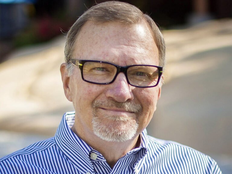 A smiling man with glasses and a blue striped button-down shirt