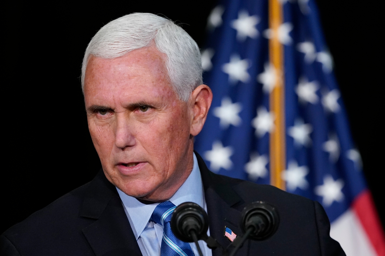 A man standing in front of a microphone with a dark suit, blue tie and shirt with an American flag behind him.