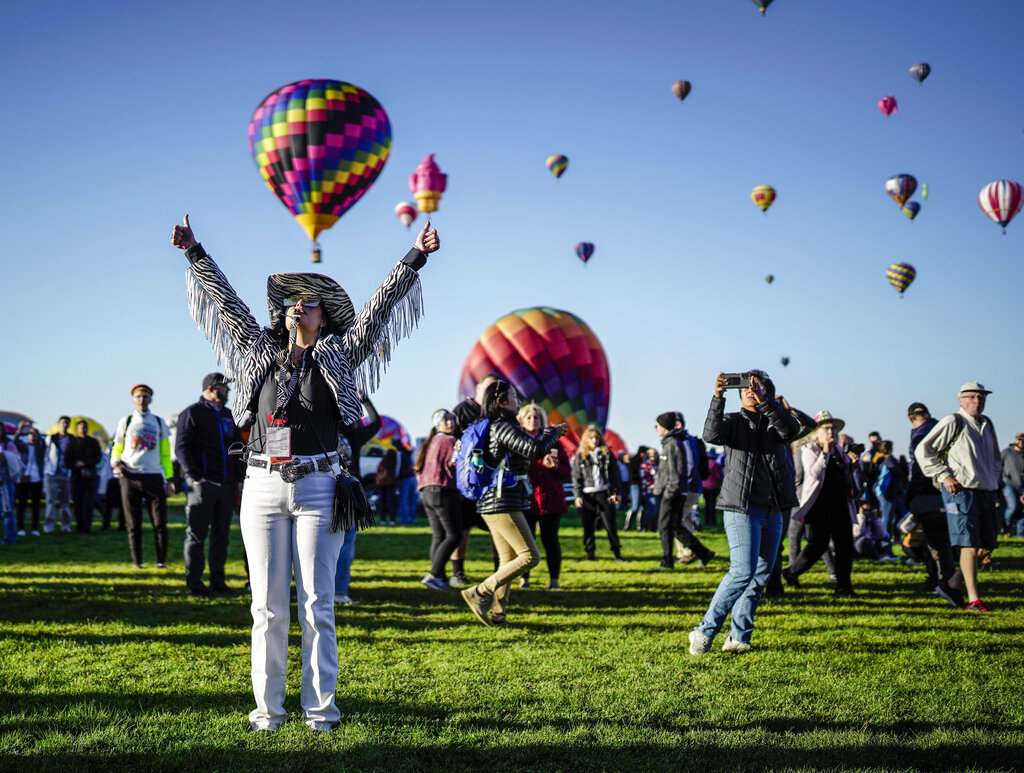 Image shows dozens of hot air balloons in the sky with spectators on the ground