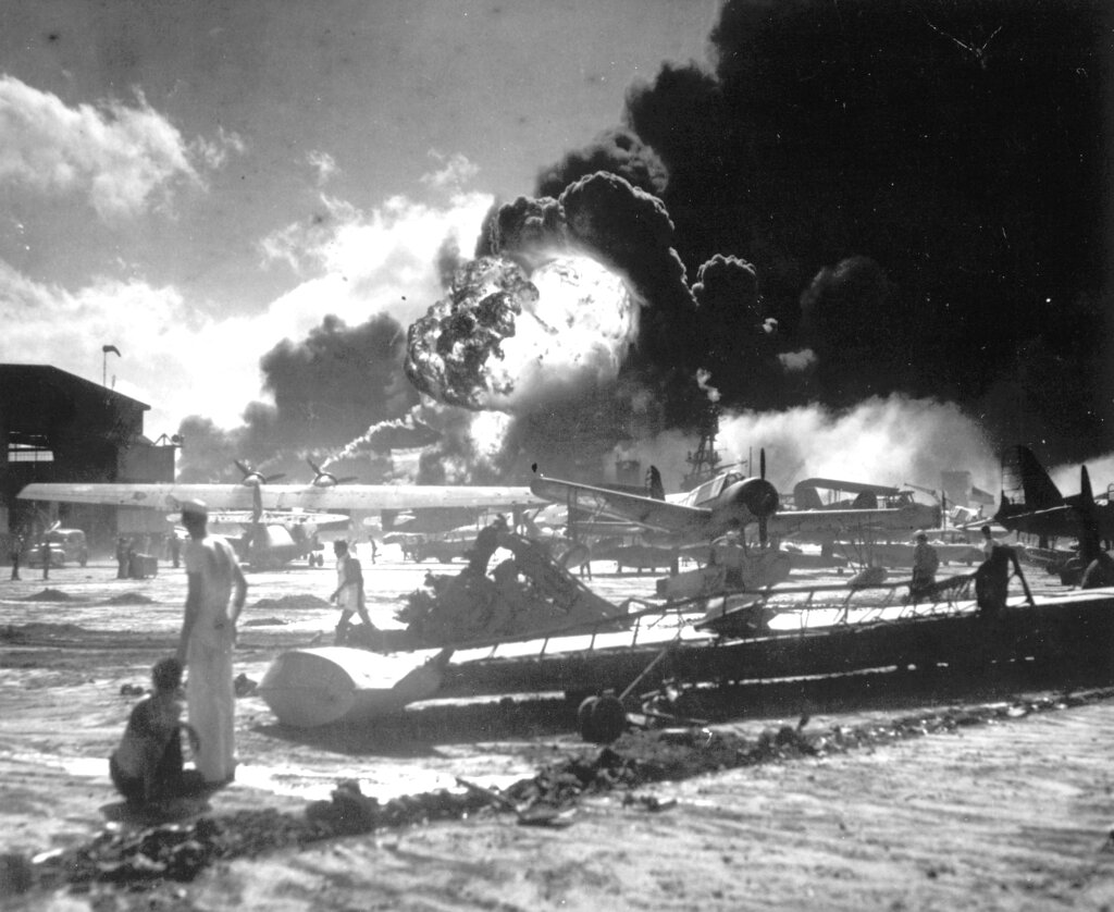 U.S. sailors stand among wrecked airplanes at Ford Island Naval Air Station as they watch the explosion of the USS Shaw in the background, during the Japanese attack on Pearl Harbor